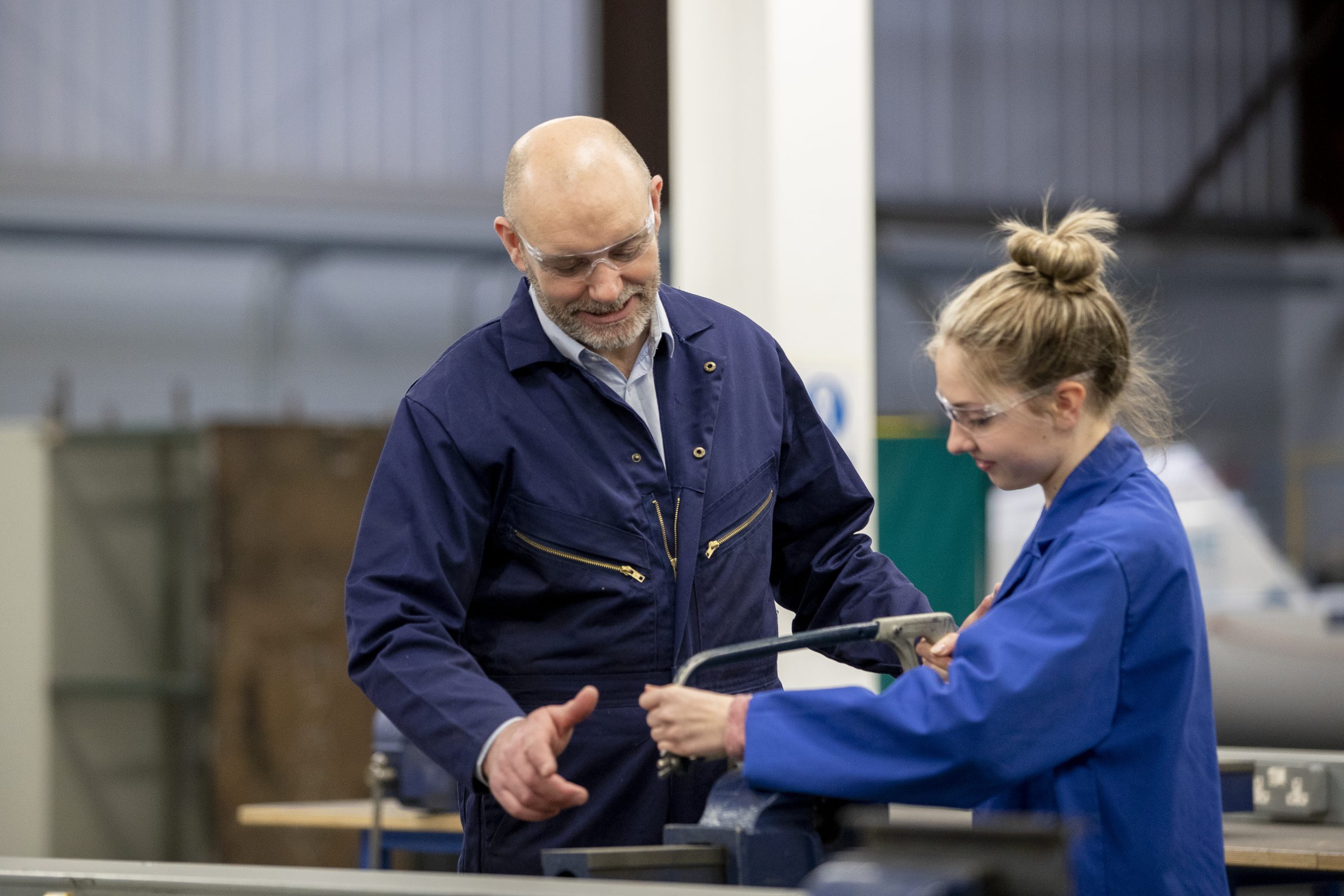 A female trainee uses a saw in an engineering workshop whilst her employer helps her.