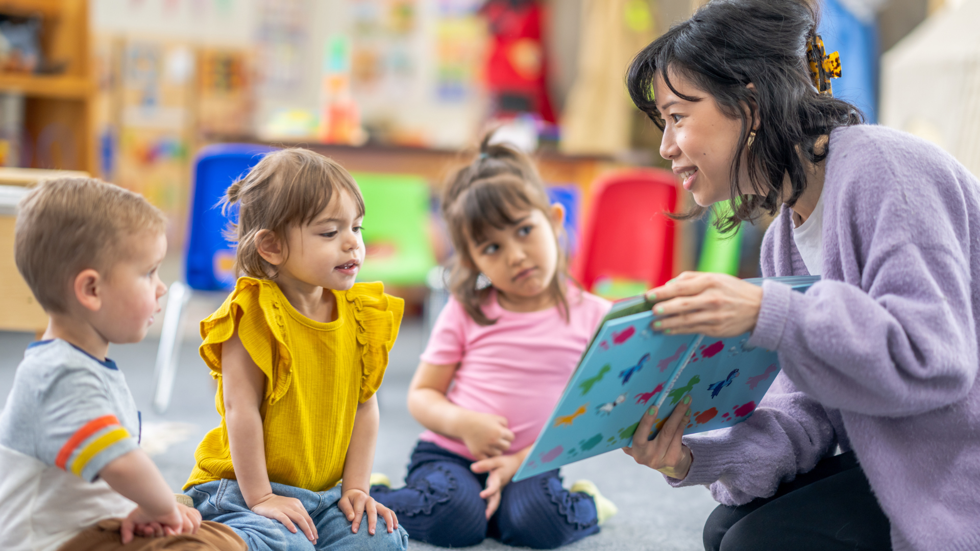 A preschool teacher sits on the floor of her classroom with a small group of students as she reads them a book. The children are each dressed casually and are focused on the story.